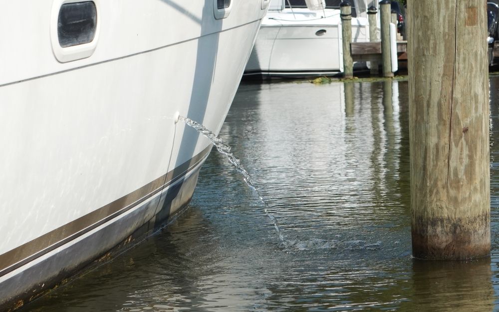 Wasser strömt von der Seite eines Bootes in einen Jachthafen, mit anderen Booten und einem Holzpfosten im Hintergrund.