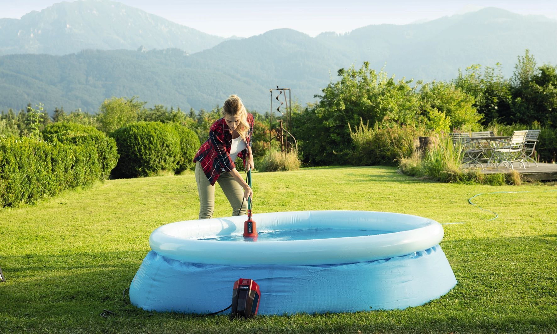 Een persoon in een geruit hemd leegt met een waterpomp op accu een blauw opblaasbaar zwembad in een met gras begroeide achtertuin met bergen op de achtergrond.