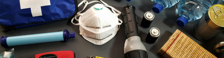 Noodpakket, waaronder een EHBO-doos, mondkapjes, zaklamp, batterijen, waterflessen, ingeblikt voedsel en een radio, op een oppervlak gerangschikt.