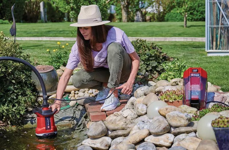 Een vrouw met een hoed verzorgt op een zonnige dag een tuinvijver met een Einhell waterpomp op accu, omringd door rotsen en groen.