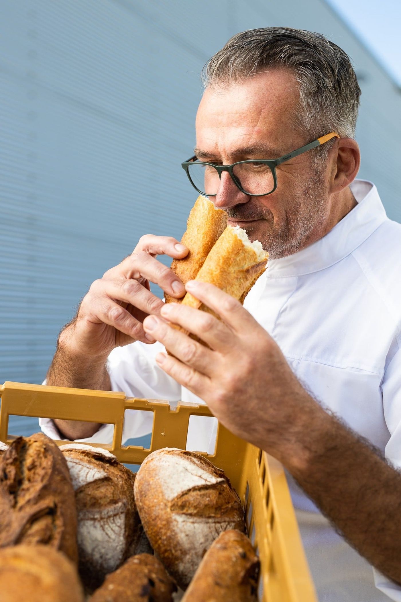 A man in glasses wearing a white coat smells fresh bread baguettes next to a basket of loaves.