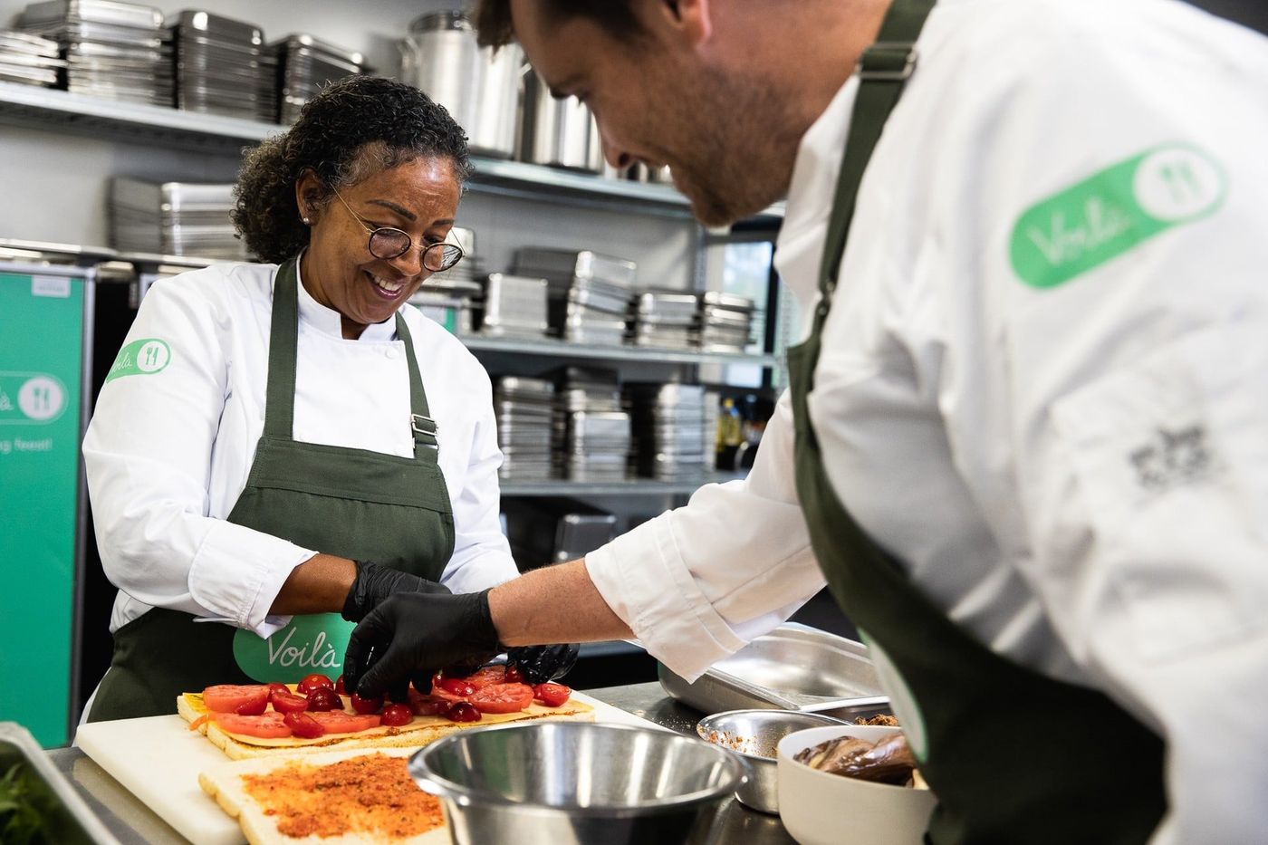 Two chefs in white uniforms and green aprons prepare food together in a professional kitchen, smiling and engaging with each other.