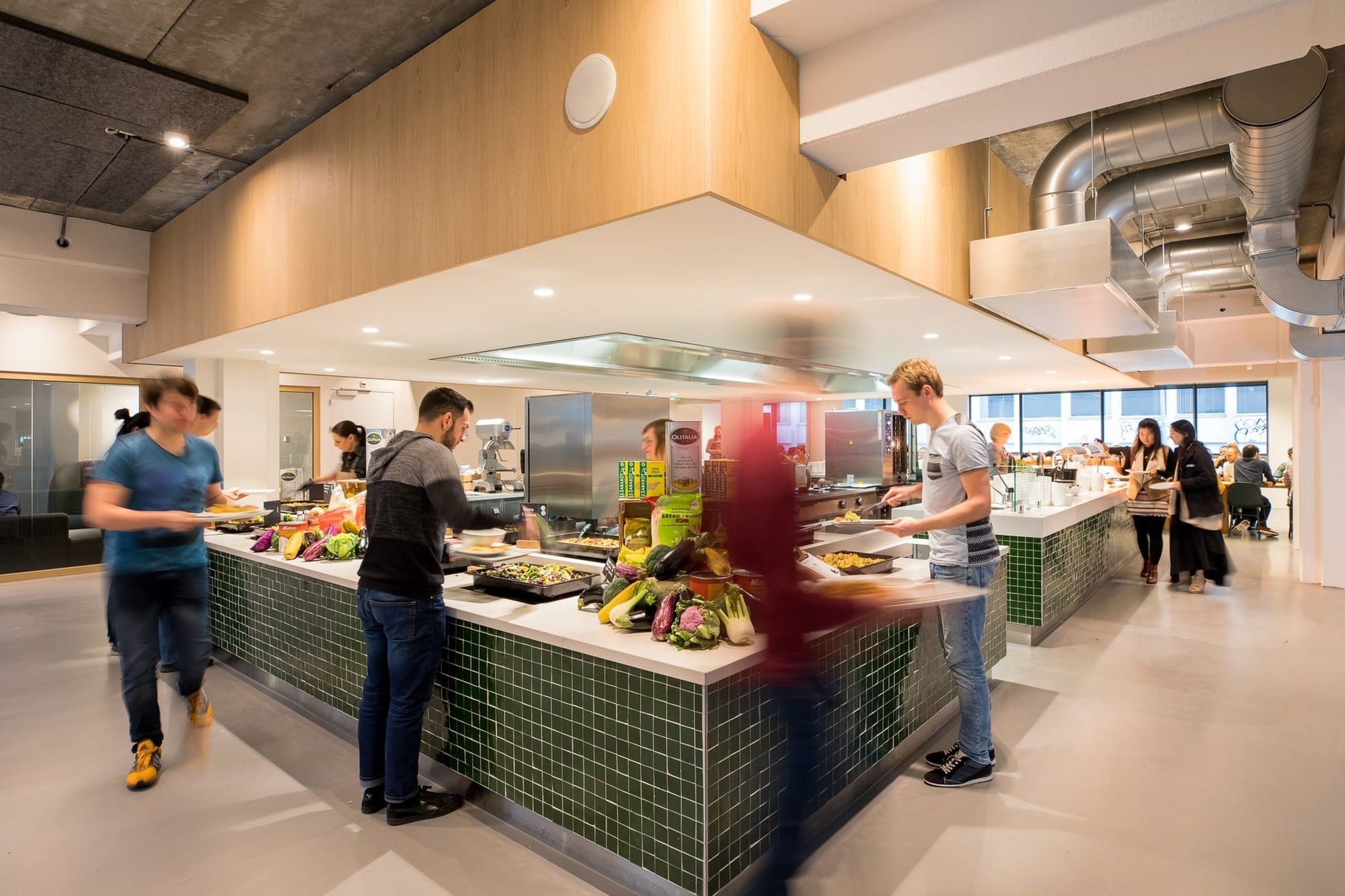 People serving themselves food at a modern cafeteria with a variety of dishes on a green-tiled counter.