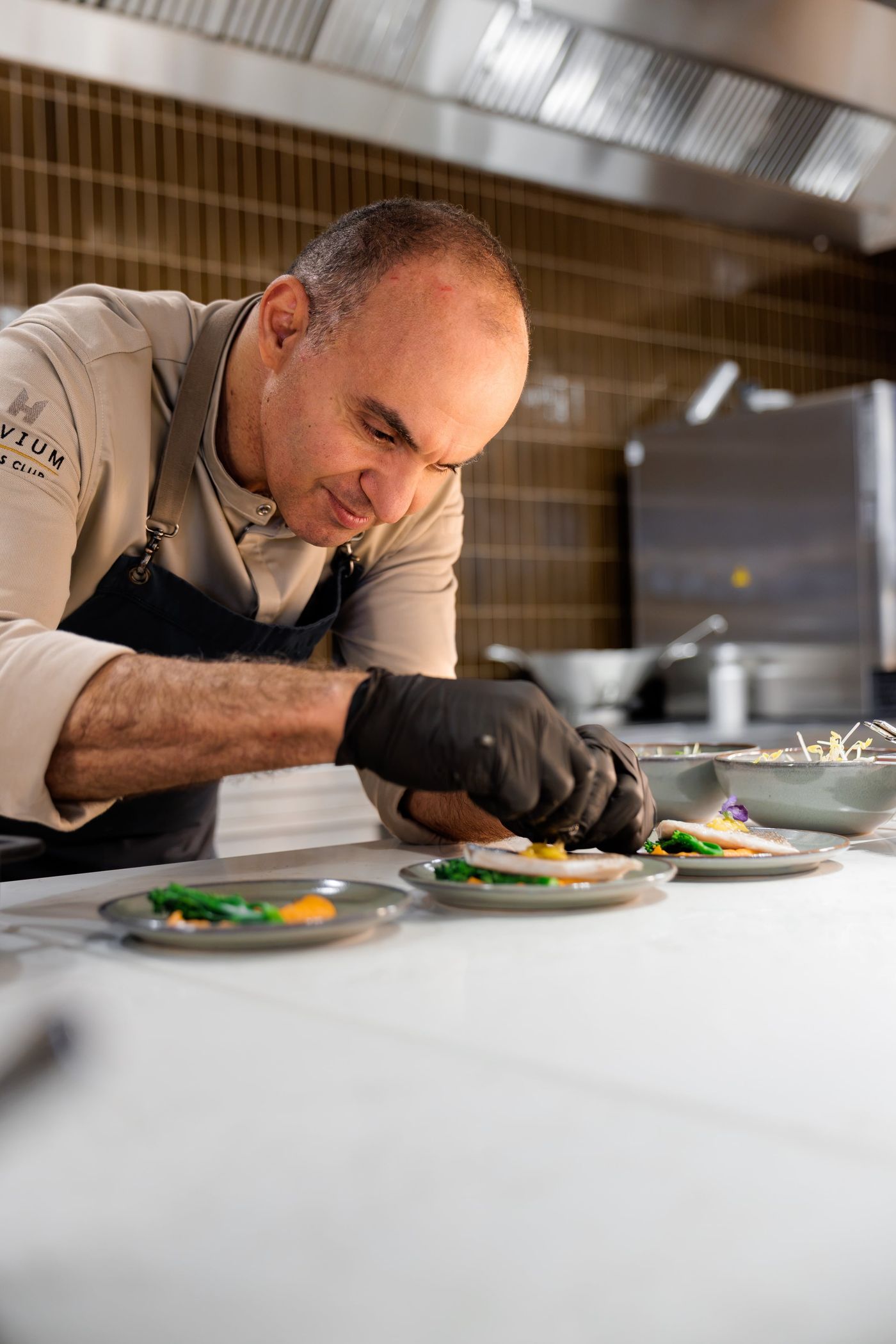 Chef in a kitchen carefully garnishing plates with colorful food using precision, wearing gloves and an apron.
