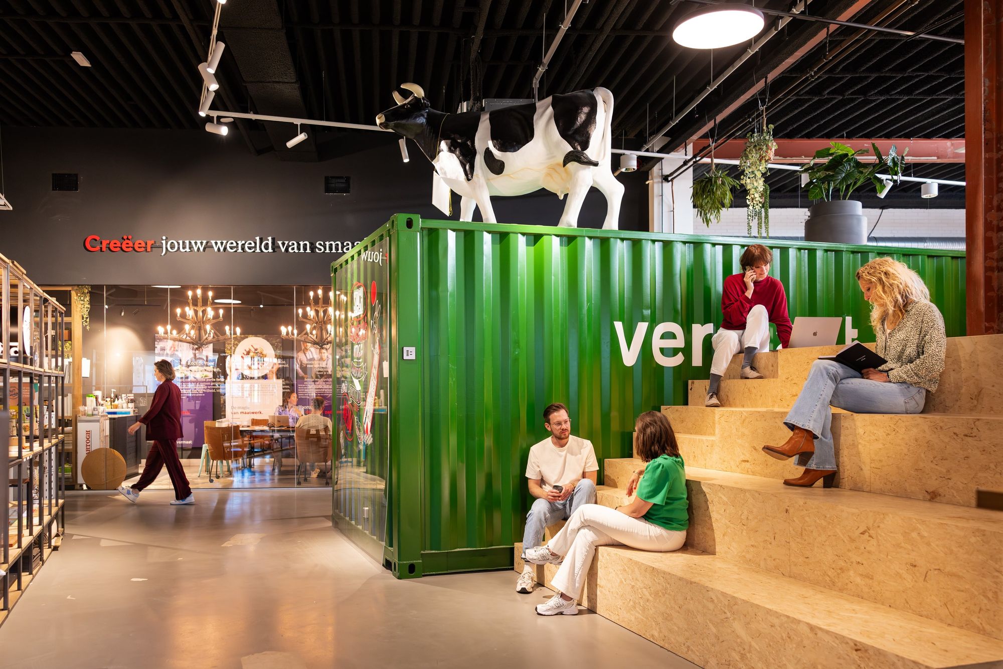 People sitting on wooden steps inside a modern space with a green shipping container and a cow statue on top.