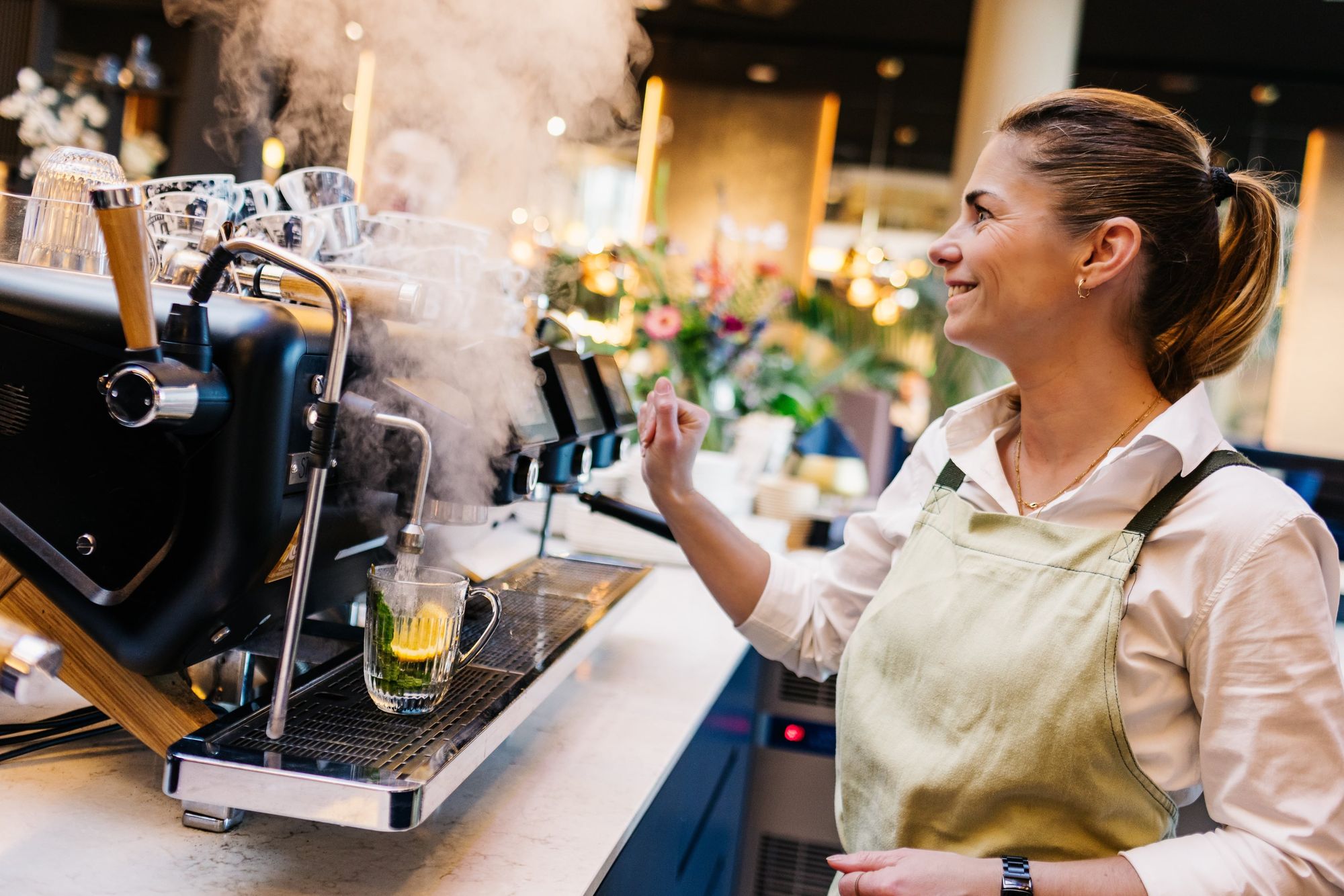 A barista in an apron smiles while steaming a beverage, with steam rising from the machine in a warmly lit café setting.