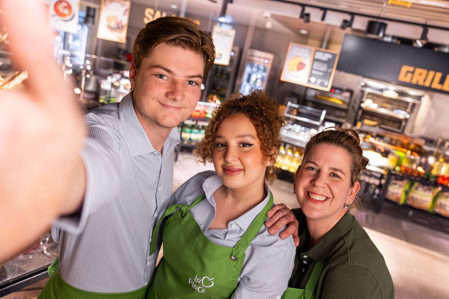 Three smiling individuals in green aprons take a selfie in a bright, modern kitchen or food preparation area.