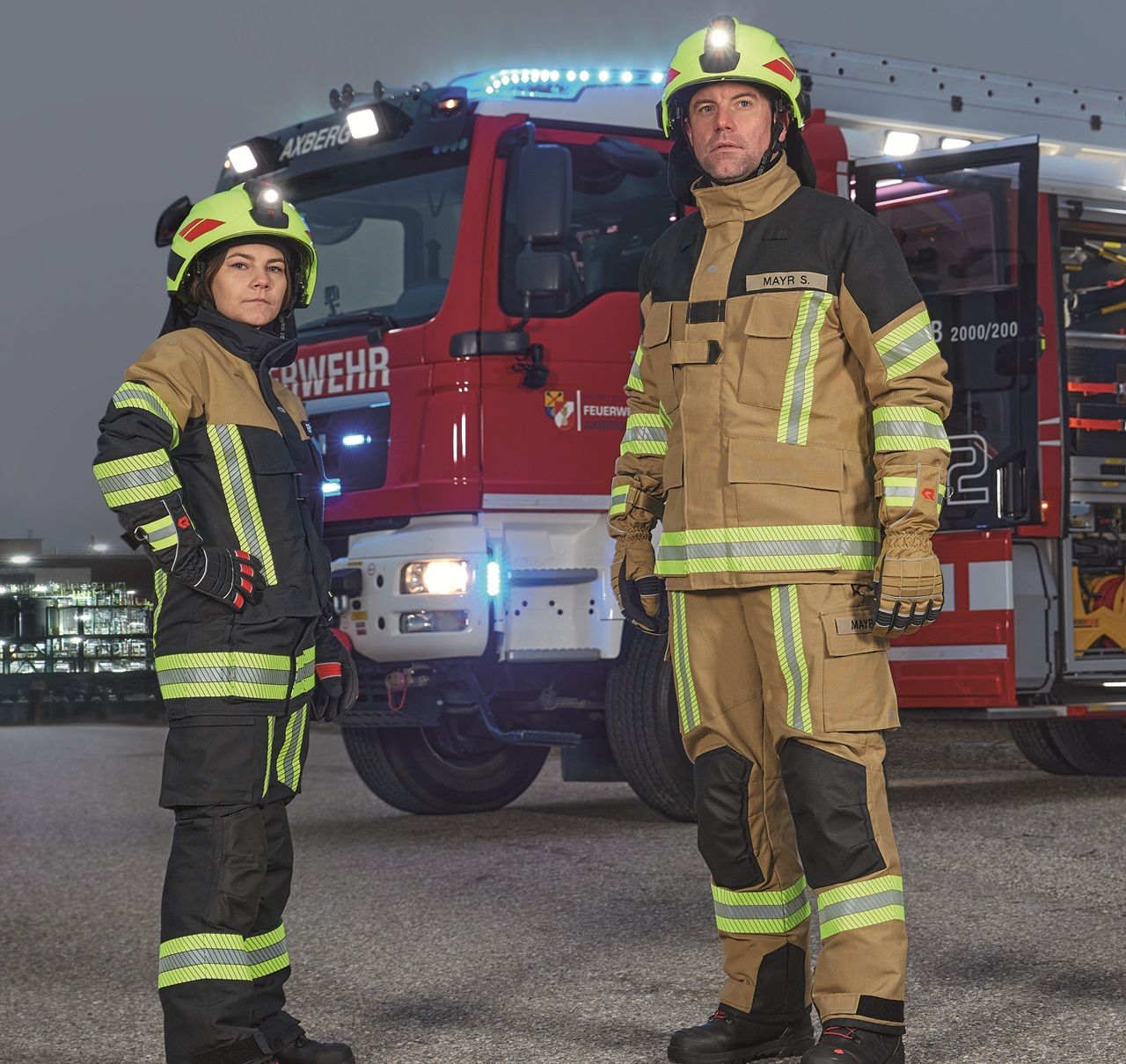 Two firefighters in protective gear stand in front of a fire truck with lights on, ready for action in an outdoor setting.