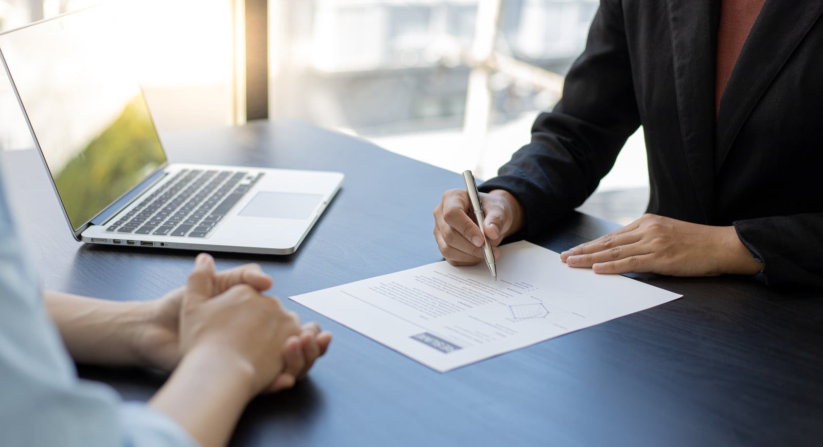 Two people at a desk with a laptop, one signing a document. Sunlight filters through a window, illuminating the workspace.