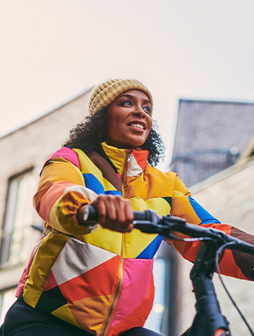 Vrouw met kleurig winterjack op een fiets, lachend en met gebouwen op de achtergrond