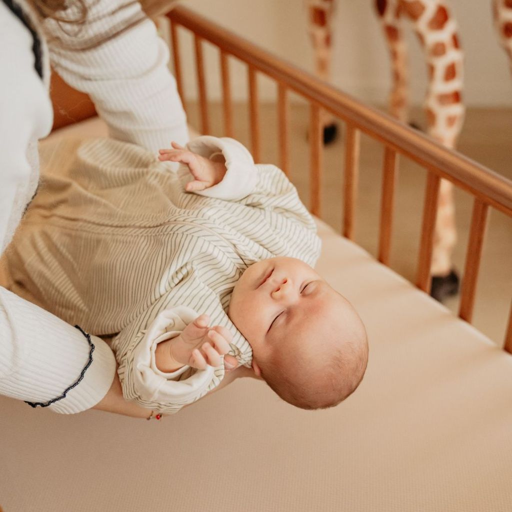 Mom holding a sleeping baby in a striped outfit over a wooden crib.