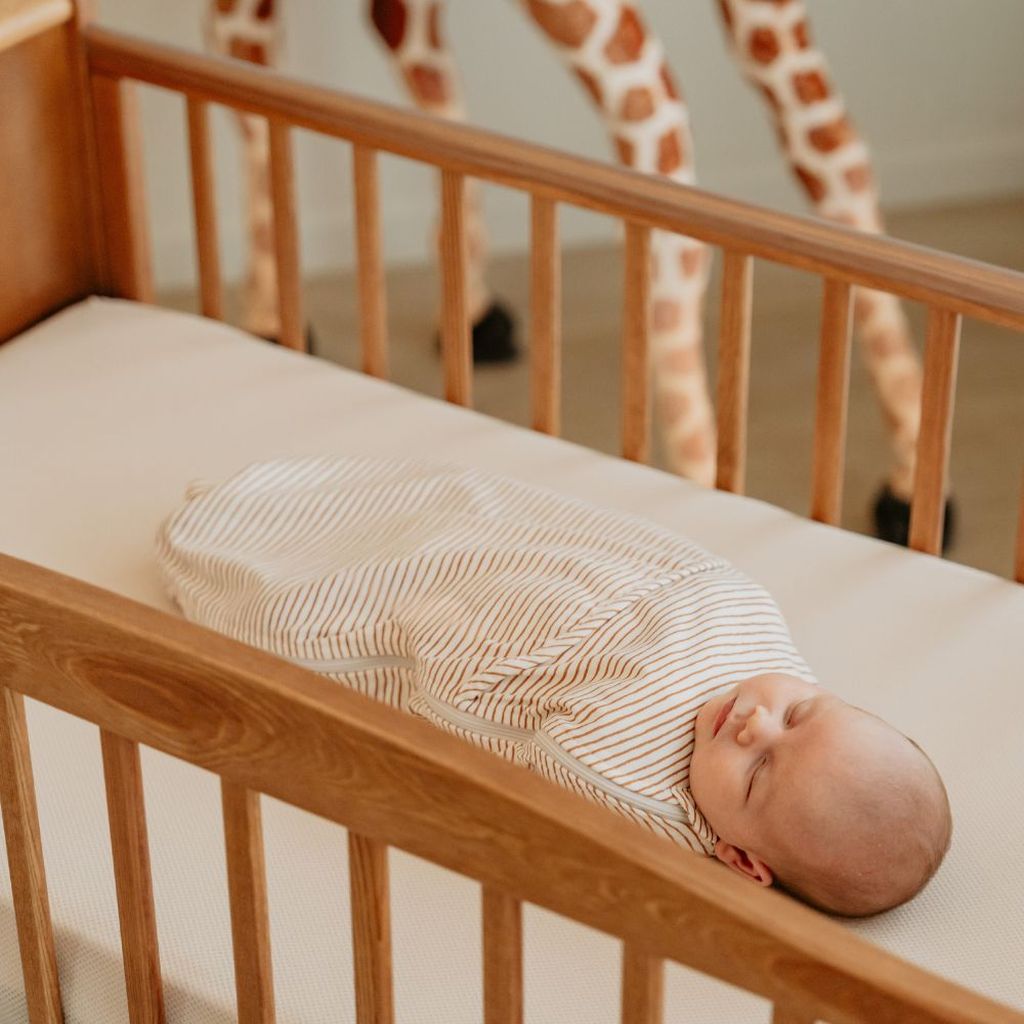 A baby wrapped in a striped swaddle sleeps peacefully in a wooden crib.