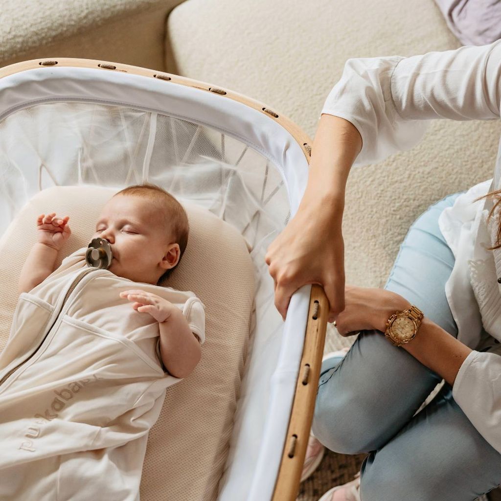 Een baby ligt in een co-sleeper. Een moeder zit naast de co-sleeper en houdt een oogje in het zijl.