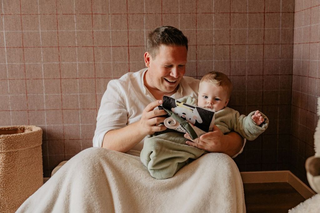 A man in a white shirt sits with a smiling baby on his lap, reading a colorful children's fabric book in a cozy room.