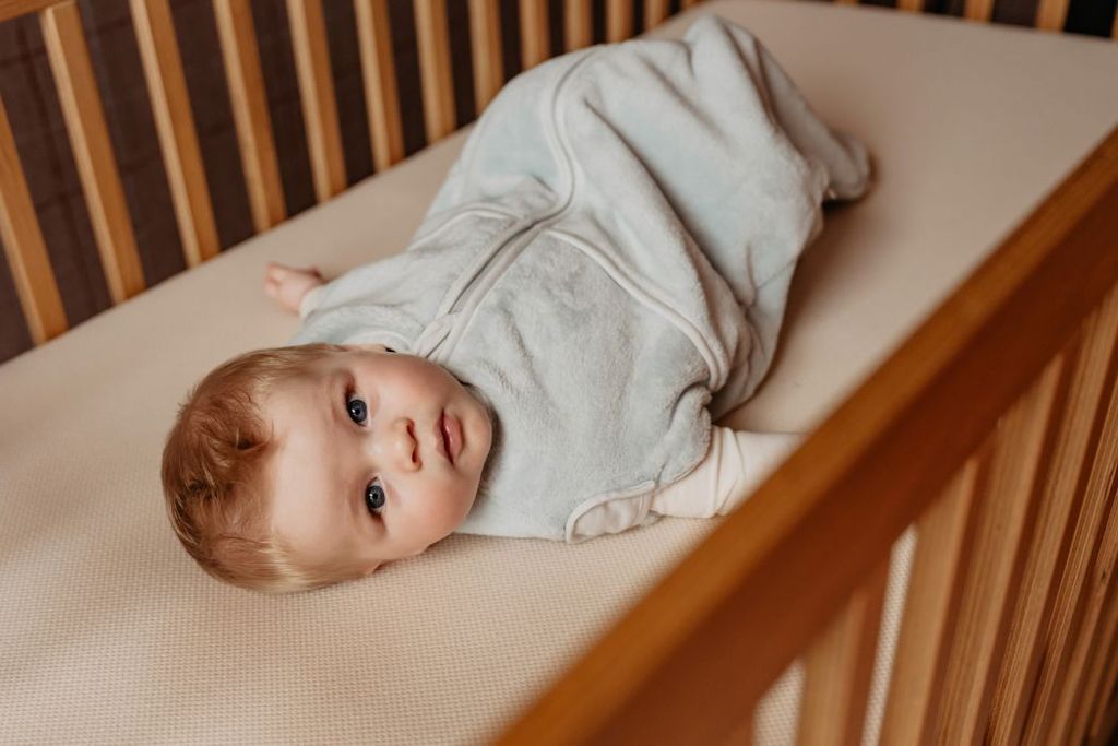 A baby in a light blue teddy Puckababy sleeping bag lies on a beige AeroSleep mattress inside a wooden crib, looking curiously at the camera.