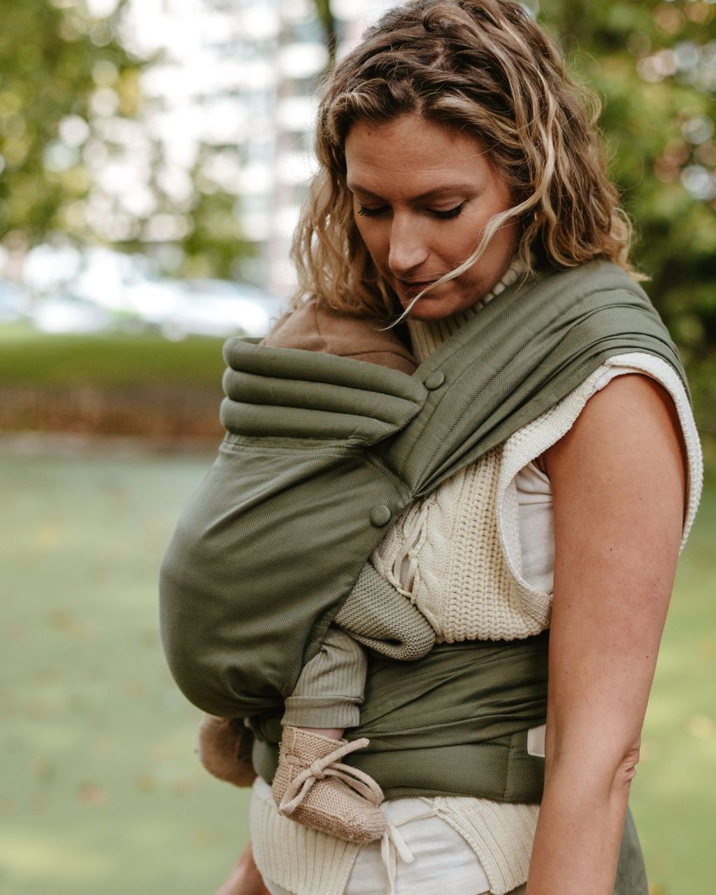 A woman in a white sweater holds a baby in a green wrap carrier outdoors, surrounded by greenery.