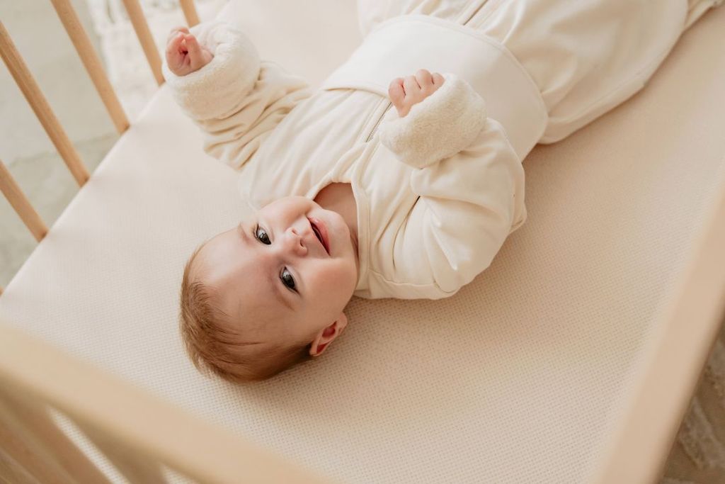 Vrolijke baby in een ledikant, gekleed in een beige slaapzak.