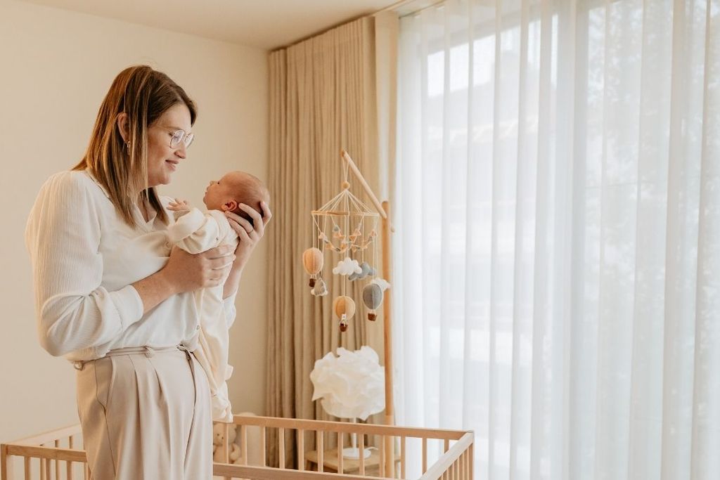 A woman gently holds a baby in a softly lit room with a crib and a mobile, near sheer curtains.