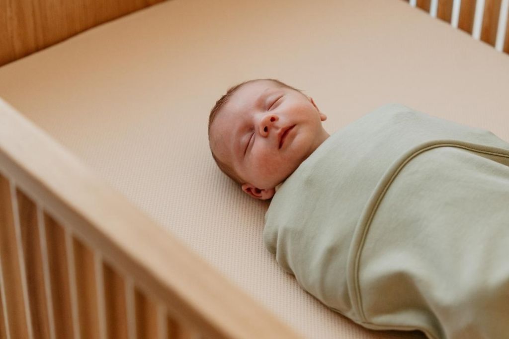 A peacefully sleeping baby wrapped in a light green swaddle lies on a beige mattress in a wooden crib.