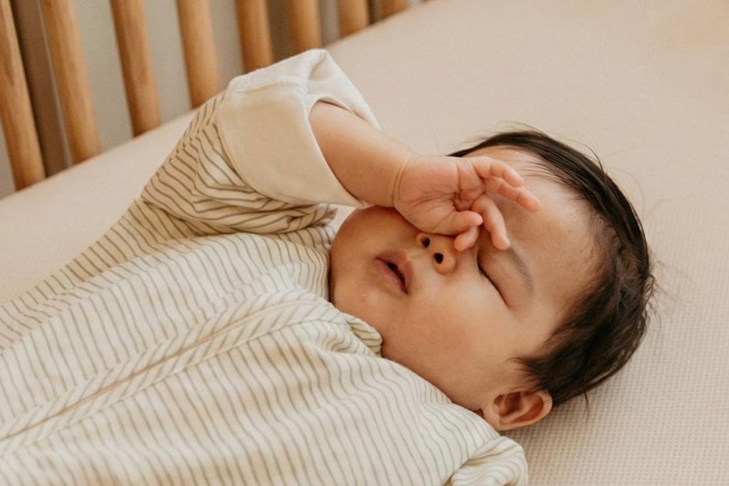 A baby in a striped onesie lying in a crib, rubbing their eye with a hand, appearing sleepy or about to fall asleep.
