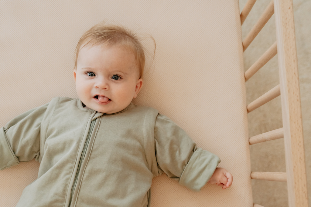 Baby lying in a crib wearing a green outfit, looking up with a slight smile and tongue out, surrounded by light bedding.