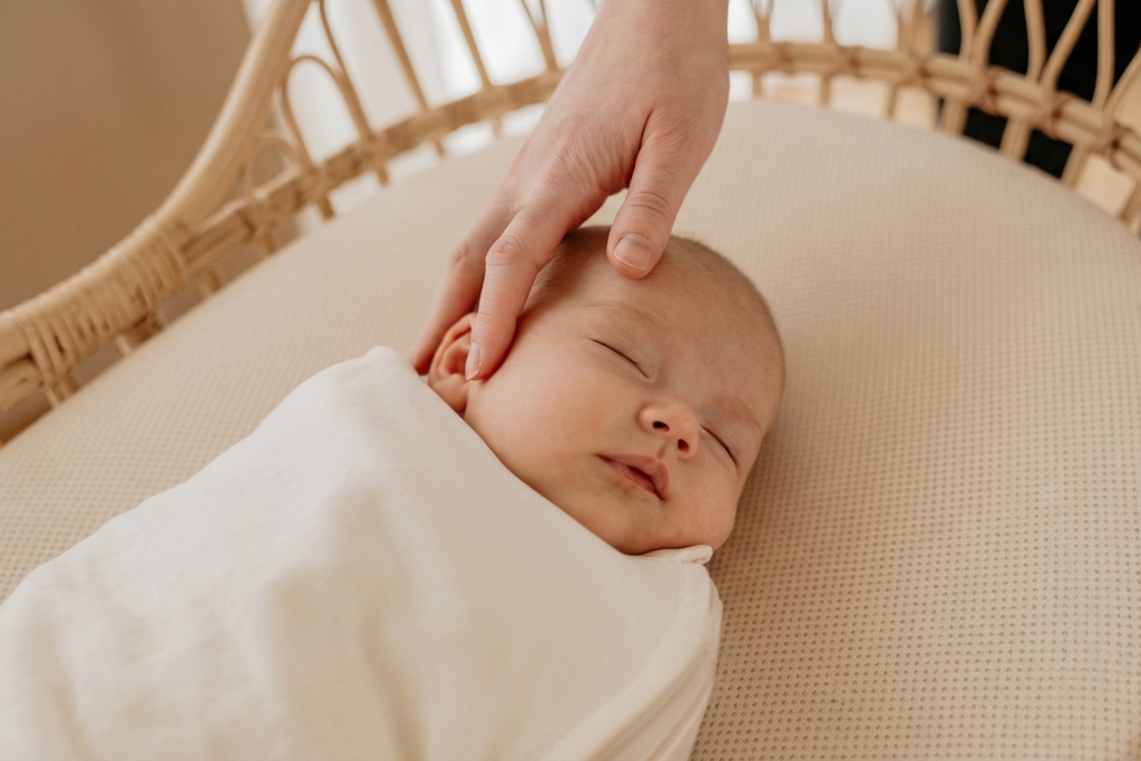 A sleeping baby wrapped in a light blanket lies in a wicker bassinet while a hand gently caresses their head.