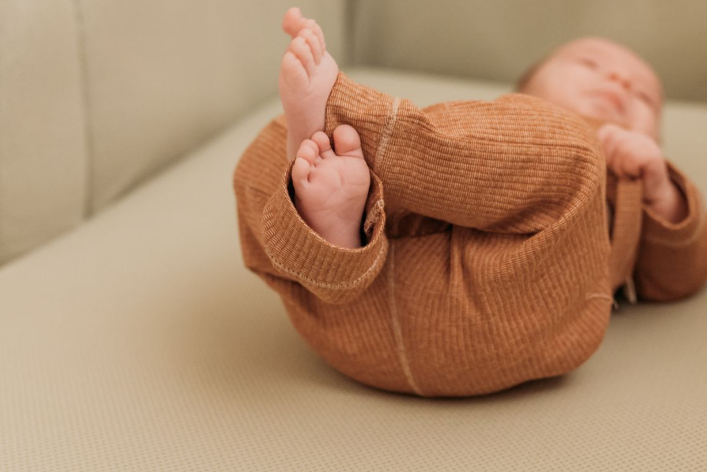 Baby in brown onesie lying on a beige surface, playfully holding their feet with both hands.