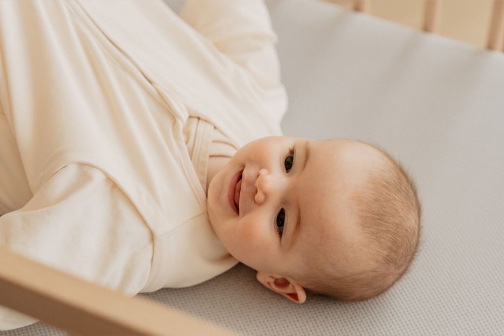 Een blije baby ligt op een AeroSleep-matraspakket met een geperforeerd hoeslaken.