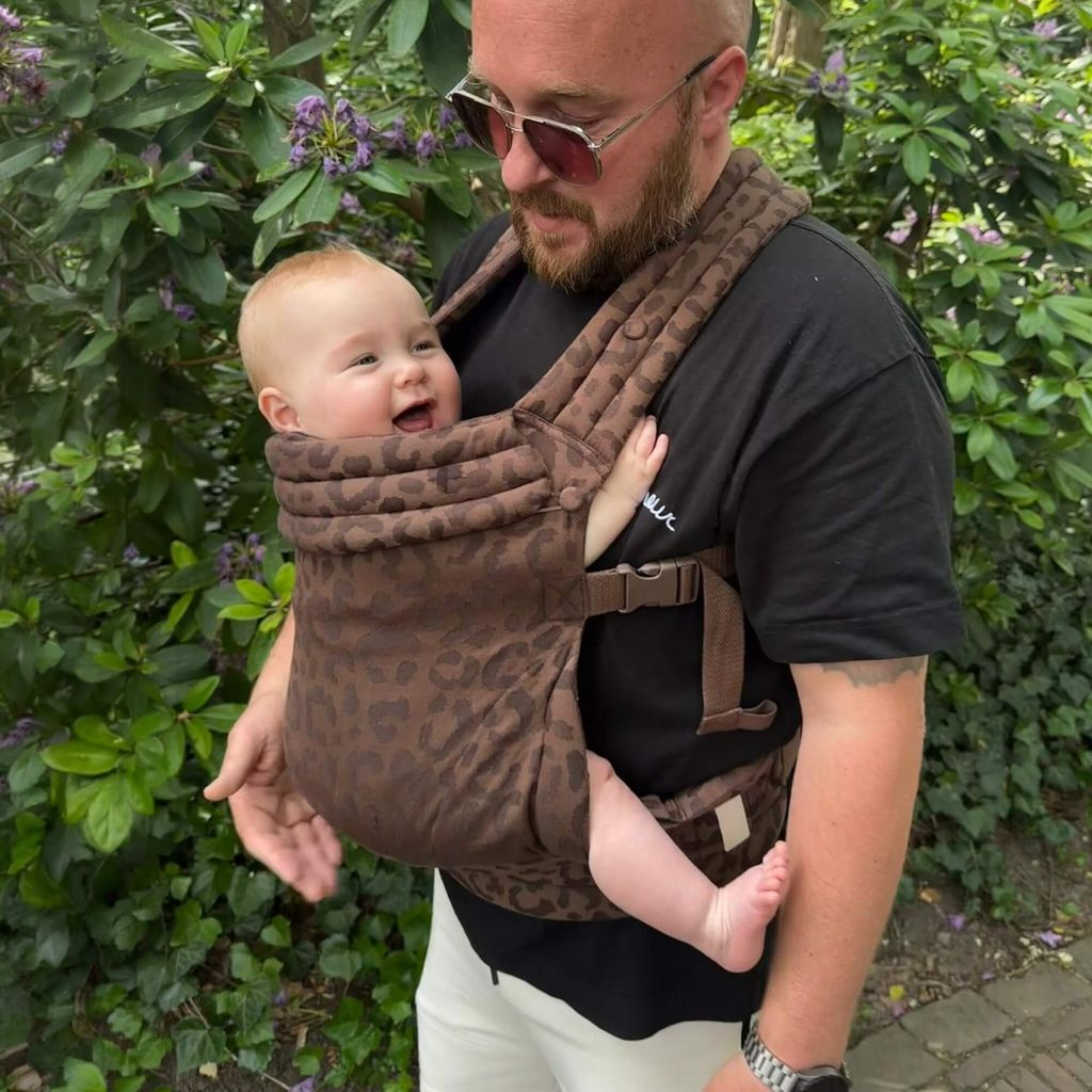 Man wearing sunglasses carries smiling baby in a brown baby carrier, standing in front of lush greenery.