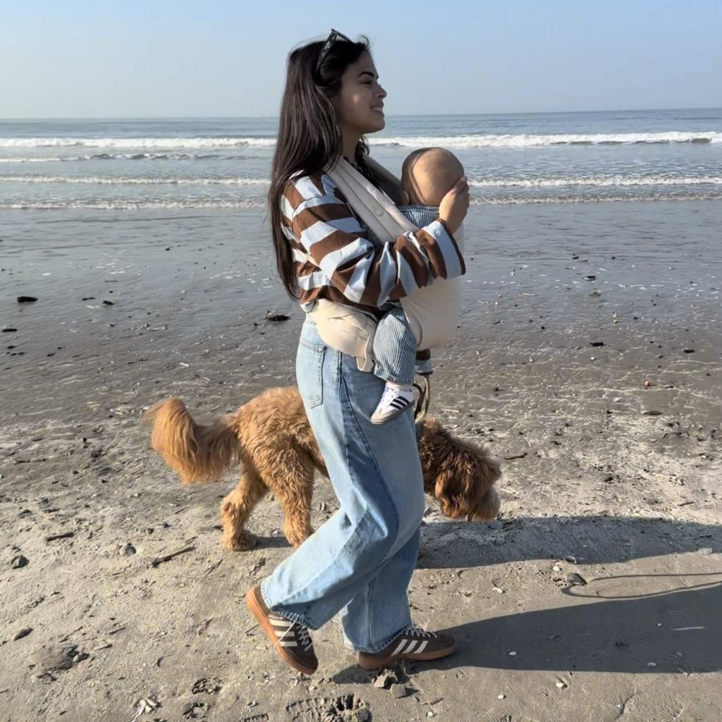 Woman walking on a beach, holding a baby in a carrier, with a golden dog beside her. Ocean and clear sky in the background.