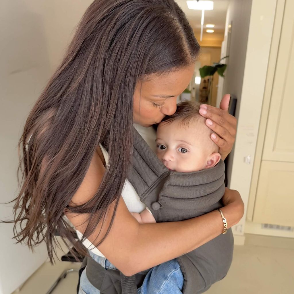 Woman gently kisses a baby's head while carrying the baby in a strap-on carrier. The setting is a bright, indoor hallway.