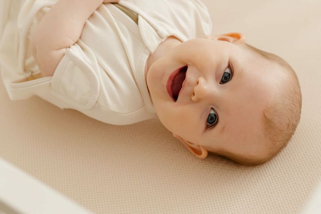 Smiling baby in a white outfit lying on a crib mattress, looking up, showing a joyful expression.
