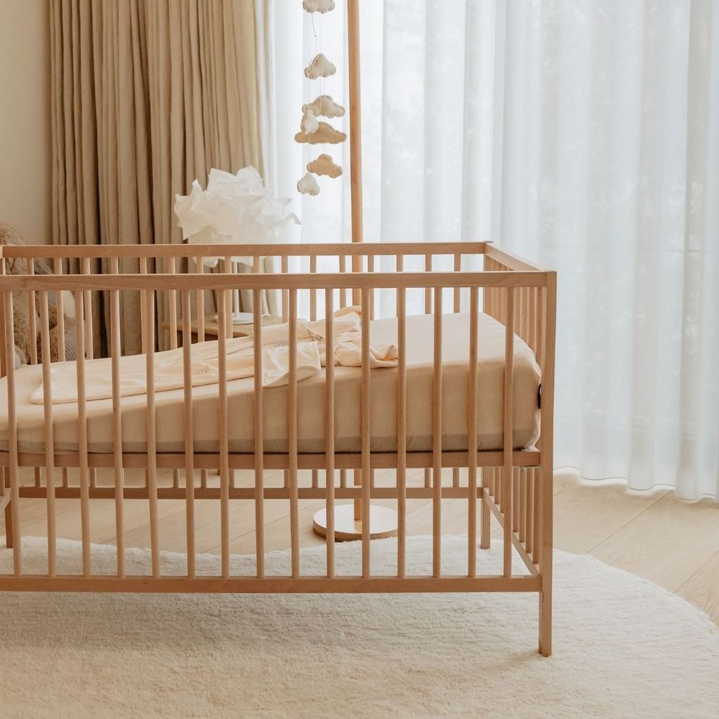 A wooden crib with beige bedding on a round cream rug. Nearby, light curtains and a white dresser create a cozy nursery atmosphere.