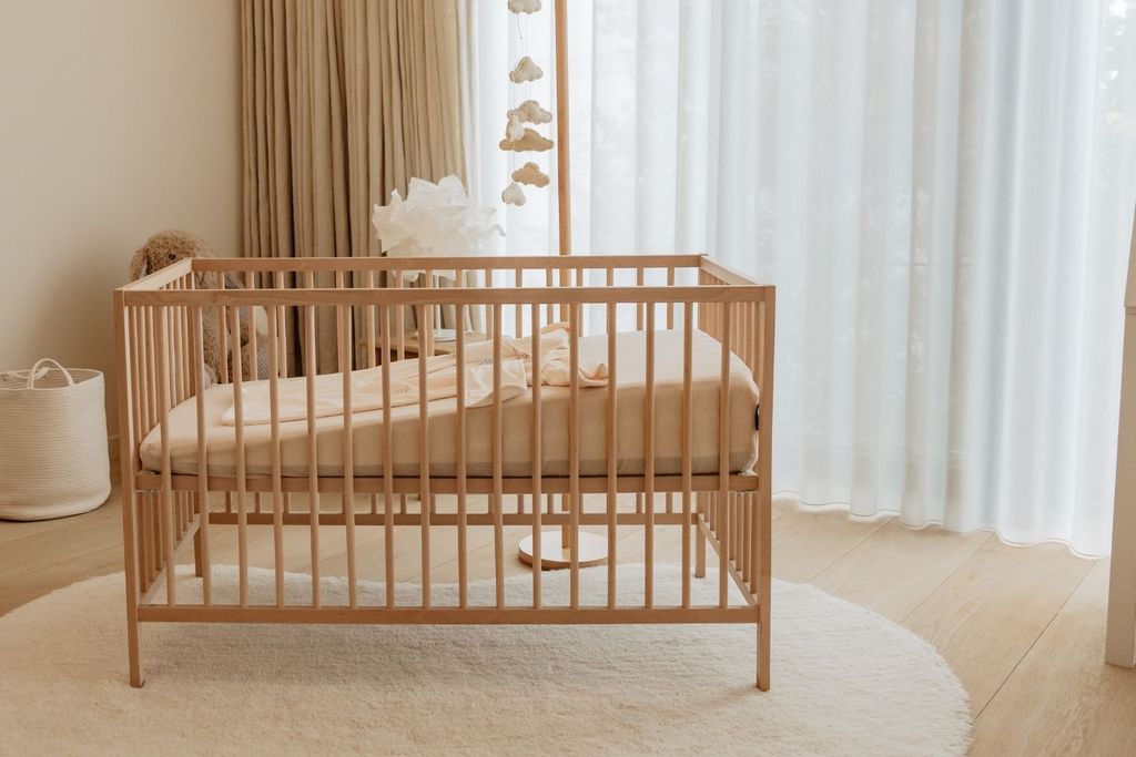 A wooden crib with beige bedding on a round cream rug. Nearby, light curtains and a white dresser create a cozy nursery atmosphere.