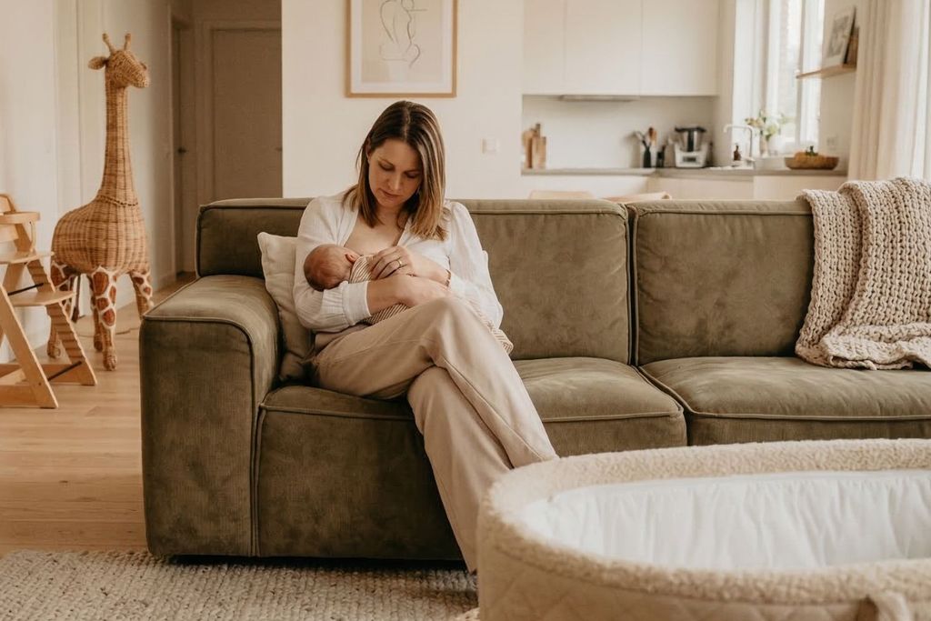 A woman sits on a green couch cradling a baby in a cozy living room with a wicker giraffe, baby bassinet, and soft lighting.