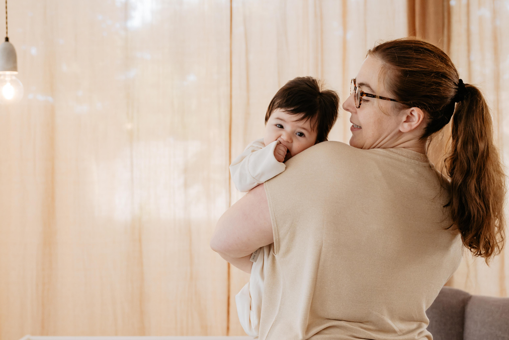 Woman holding a baby in a cozy room with beige curtains and soft lighting in the background.