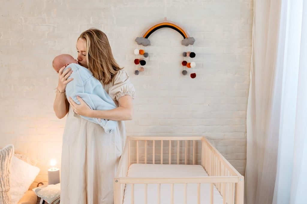 A woman gently kisses a baby wrapped in a blue blanket, standing beside a crib in a softly lit room with a rainbow decoration on the wall.