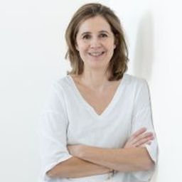 A woman with shoulder-length brown hair, wearing a white blouse, stands with arms crossed against a white background, smiling at the camera.