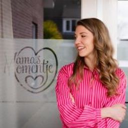 Woman in a pink striped shirt smiles while standing outside near a glass door with a heart logo and "Mamas Momentje" written on it.