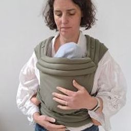 Woman in a white blouse cradles a baby in a green baby carrier, standing against a plain white background.