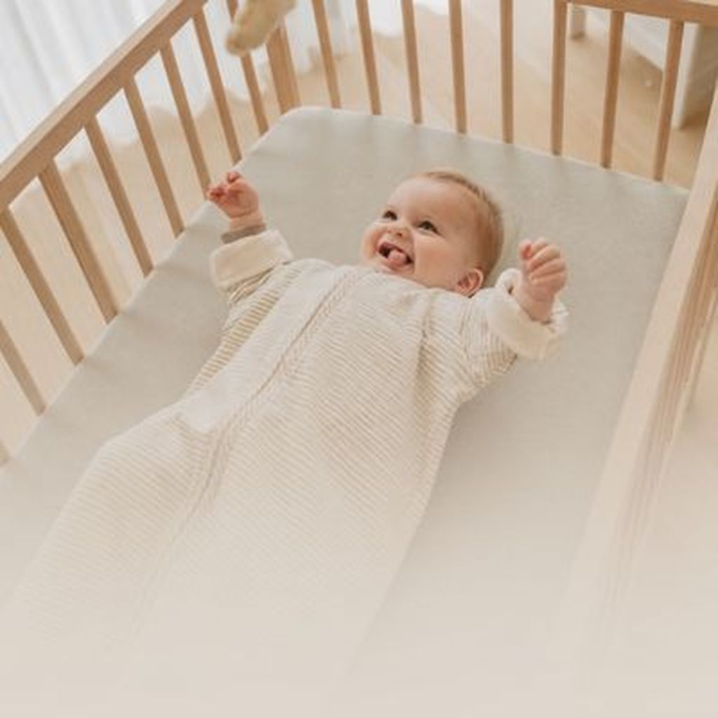 A smiling baby in a beige sleep sack lies in a wooden crib, arms raised, with soft natural light streaming in through a window.