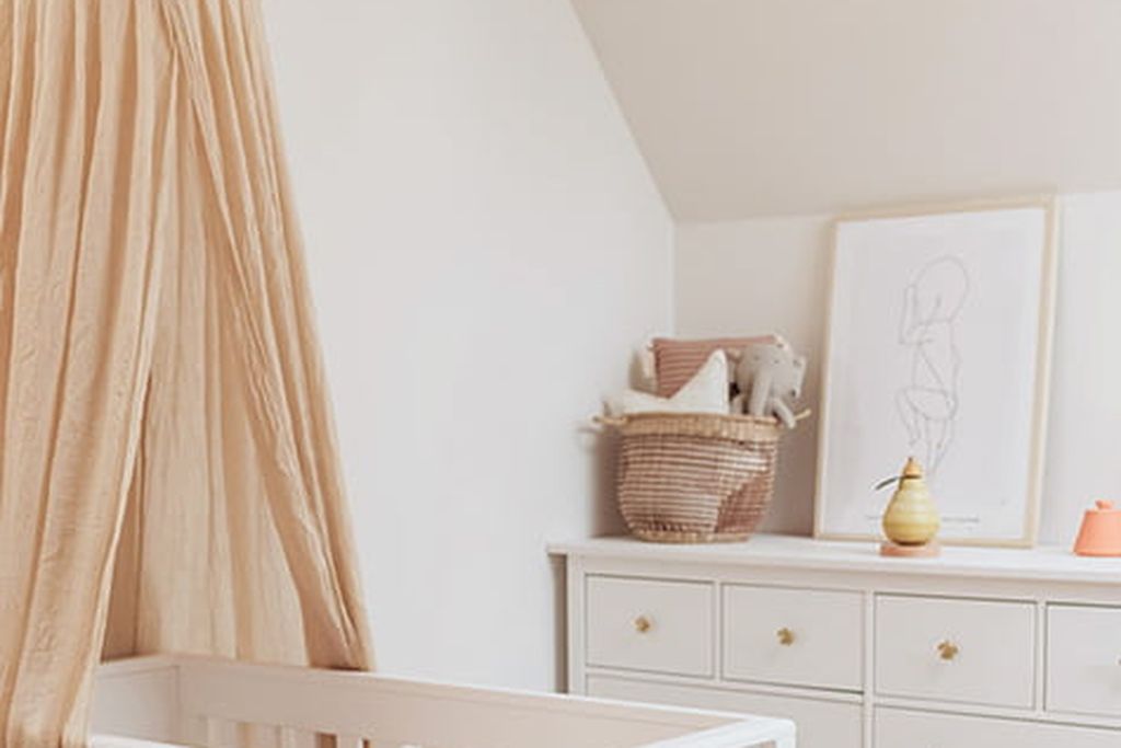 Ein Kinderzimmer mit einem weißen Kinderbett, einem beigen Bettvorhang, einer Kommode mit Deko