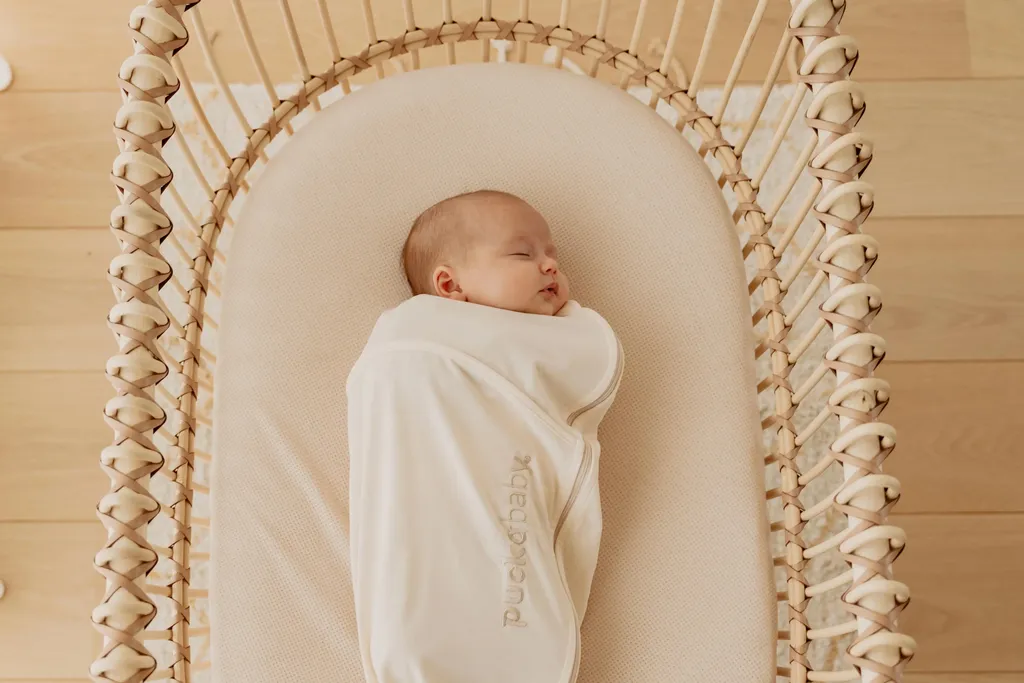 Een slapende baby, gekleed in een zachte, crèmekleurige inbakerzak, liggend op een AeroSleep hoeslaken in een gevlochten wiegje op een lichte houten vloer.