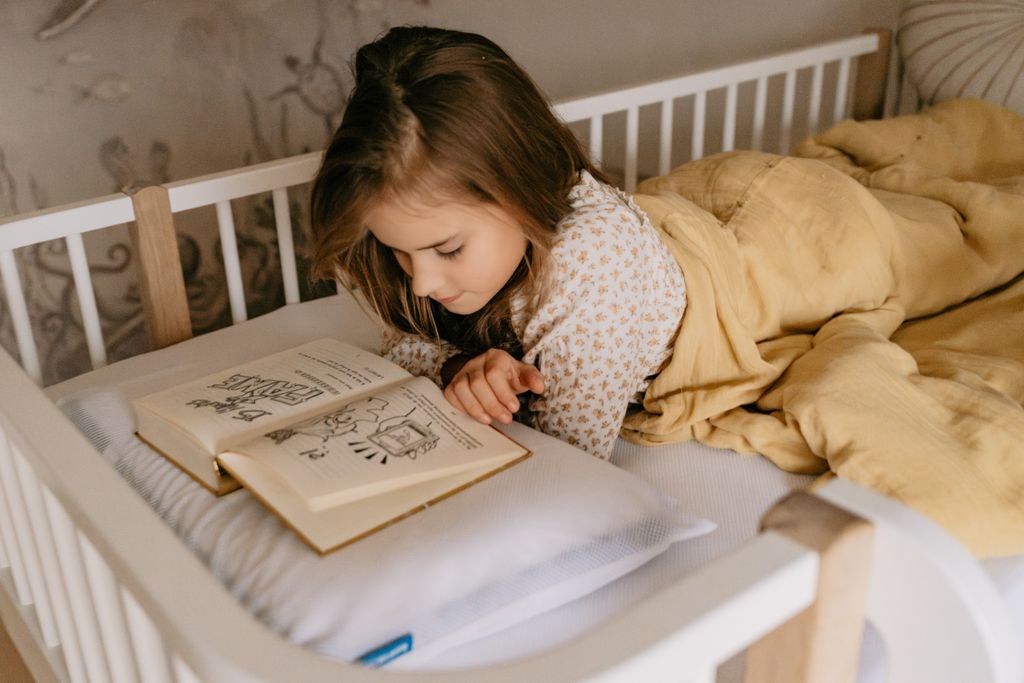 A young girl in pajamas, lying on a bed and reading a book