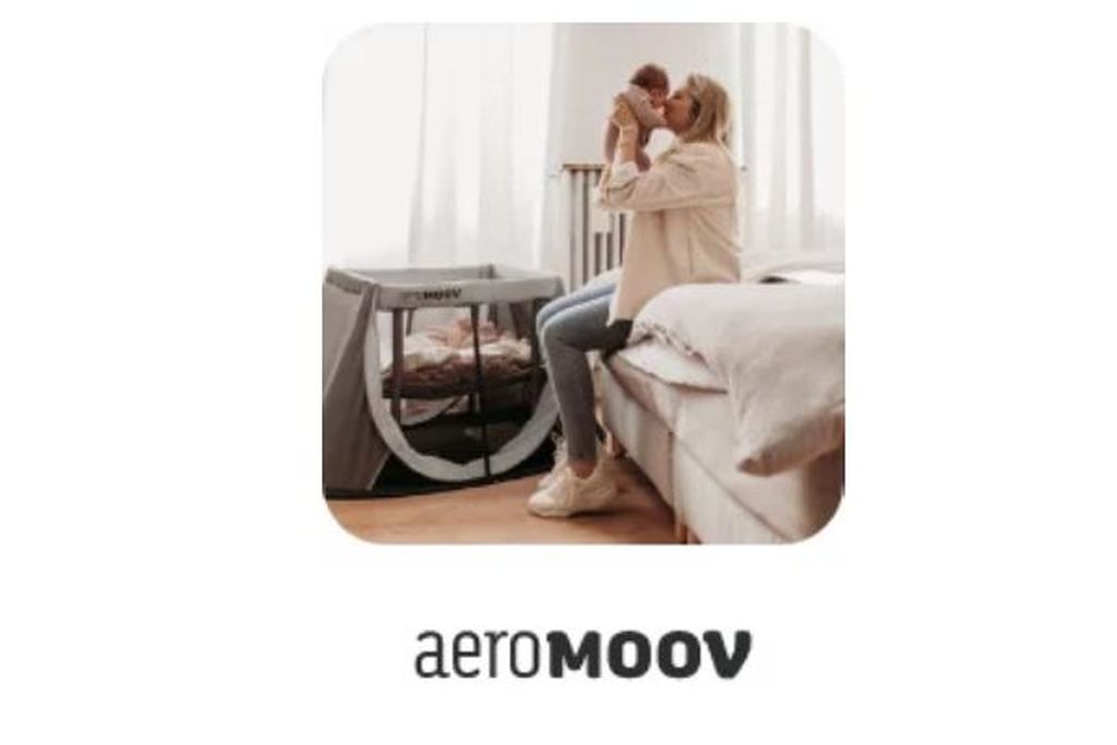 Eine Frau sitzt auf einem Bett, hält ein Baby im Arm und küsst es, neben ihr steht ein AeroMoov-Reisebett