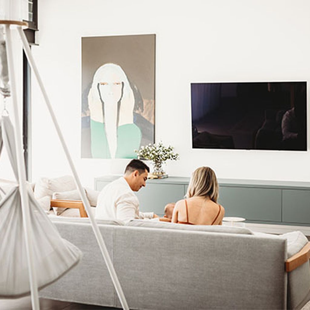 Ein Paar sitzt auf einem modernen grauen Sofa. Hinter ihnen eine NONOMO Federwiege mit Tipi Gestell