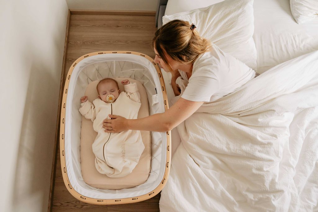 A woman gently places a sleeping baby in a crib. The baby has a pacifier and a sleeping bag.