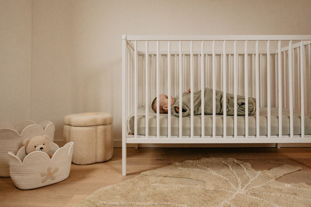 A baby in a green sleeping bag lies in a white crib in a cozy nursery