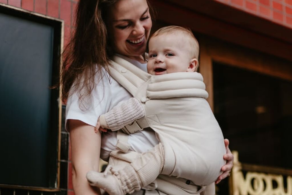 A smiling woman holds a baby in a beige carrier, standing outside near a brick wall. The baby is looking at the camera, wearing matching mittens and booties.