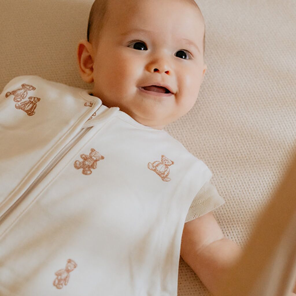 Glimlachende baby liggend op een zacht oppervlak, gekleed in een Puckababy slaapzak met Teddy Bears-print. De baby kijkt omhoog met een vrolijke uitdrukking.